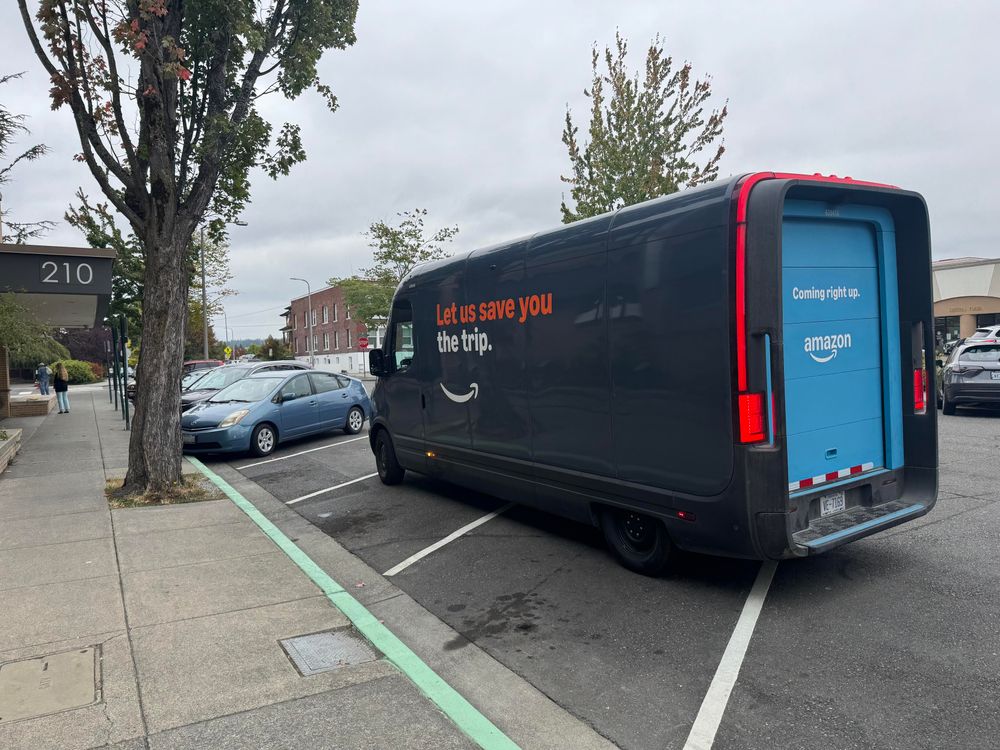 A blue Amazon delivery van parked across four parking spaces. The spaces are marked with. Green curb, indicating the free 10 minute spots at the library.