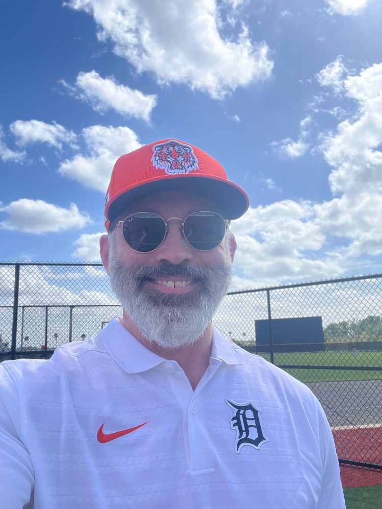 A selfie of me on a sunny day wearing a Detroit Tigers Old English D white polo and the 2025 Orange spring training cap. 