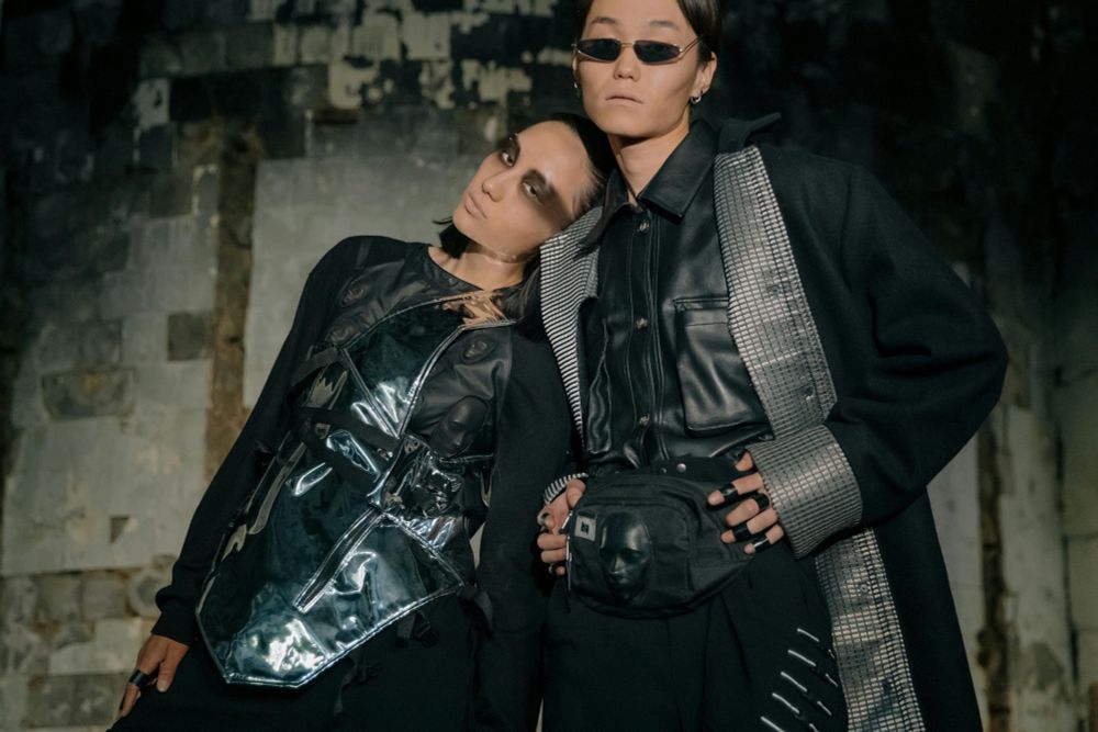 The coolest couple on earth stand in futuristic garb all in black and grey and vague shine. The slightly more masc one is wearing narrow sunglasses that coordinate perfectly with their razor-sharp cheekbones; they also have a fanny pack that manages to be cool. The slightly more femme one has Mad Max raccon eye makeup and is leaning languidly on the other's shoulder. Both assault the camera with their gaze, daring you to challenge their right to perfect gay bliss in the post-apocalyptic future