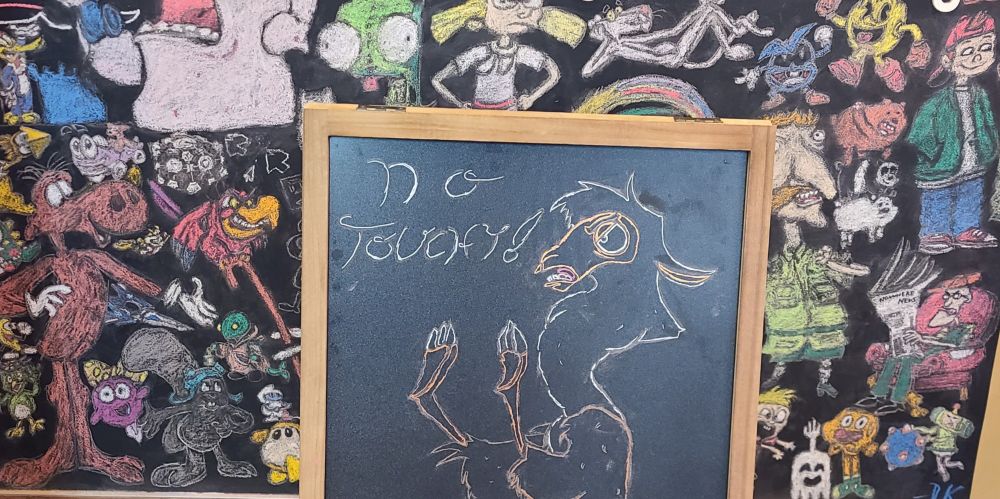 A drawing of Llama Kuzco from The Emperor's New Groove, positioned in front of a chalkboard covered in colorful drawings, with the text "NO TOUCHY!"