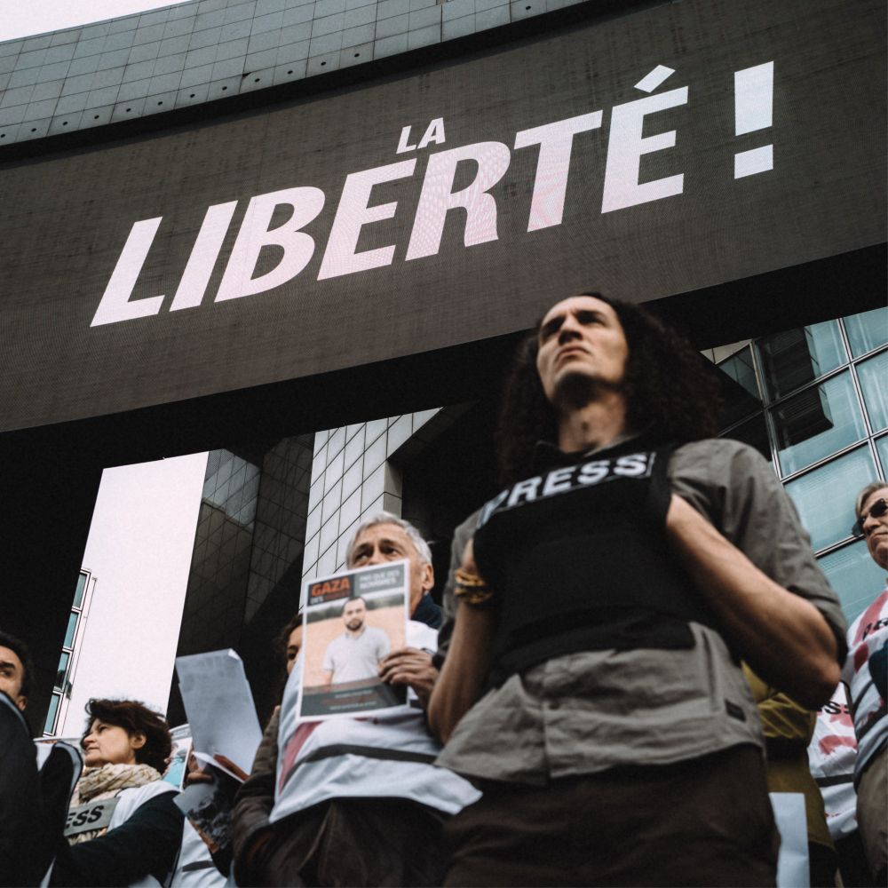 Des journalistes rassemblés sous un immense écran affichant "LA LIBERTÉ !" ; certains portent des gilets de protection presse, d’autres tiennent des portraits de confrères palestiniens tués à Gaza.