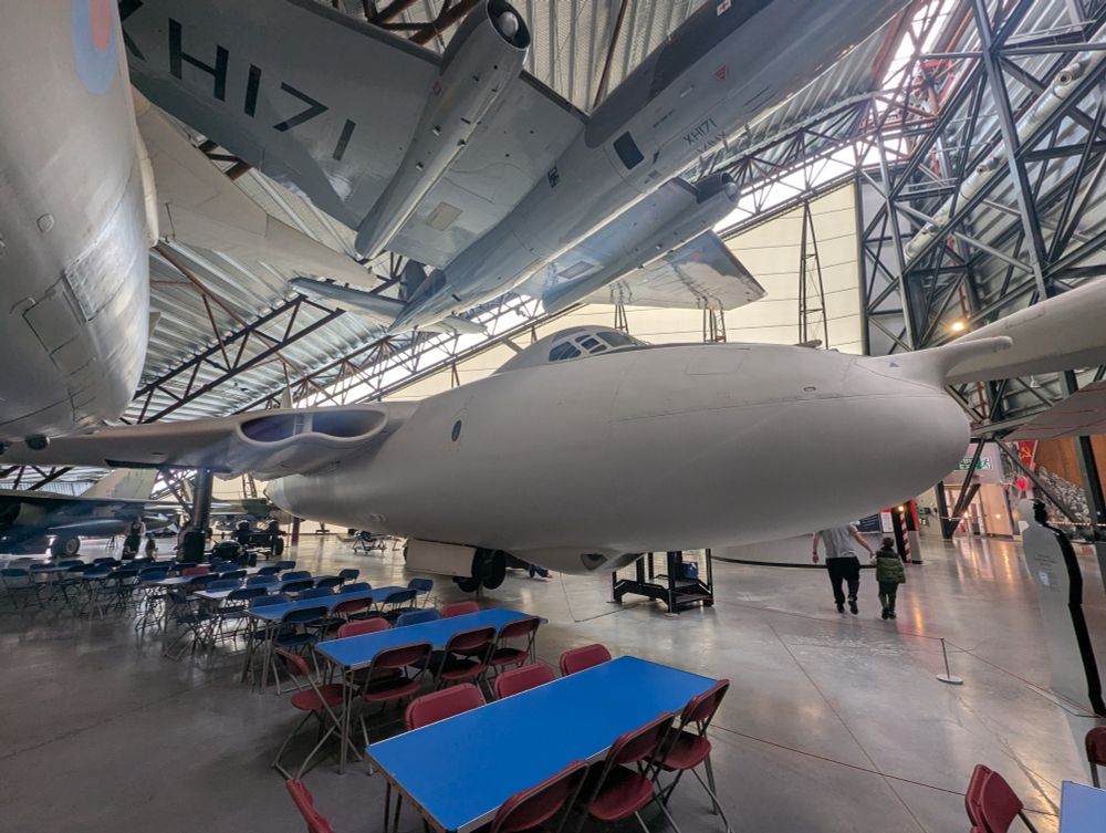 A Vickers Valiant, in white paint, in a large museum hall. Suspended over it is an English Electric Canberra. In the foreground are blue tables and red chairs.