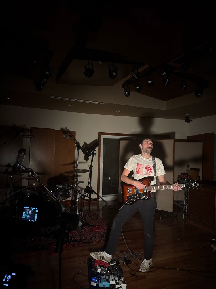 liam standing playing guitar in a recording studio with intense lighting 