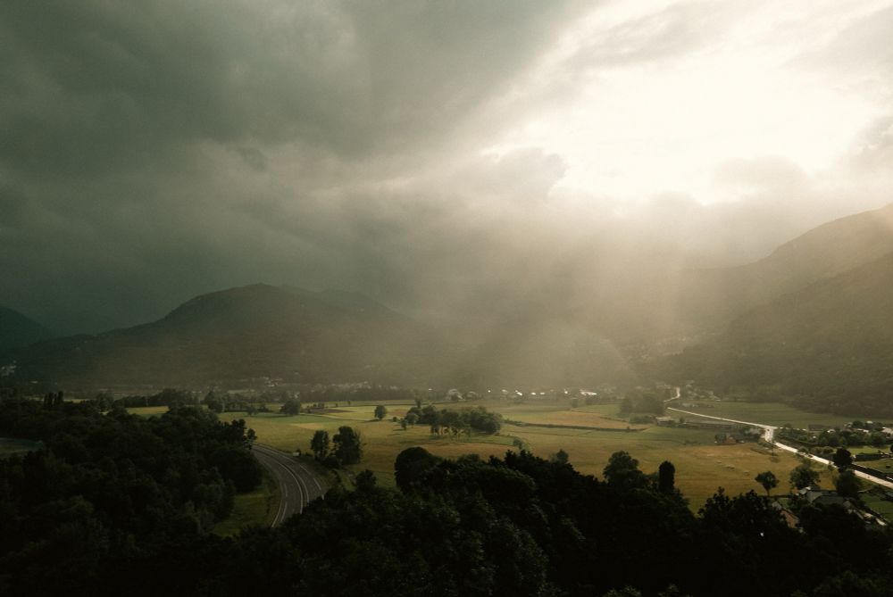 Une vue sur la vallée des Gaves dans les Pyrénées, en hauteur avec un temps orageux, c’est une vue sur des champs et les montagnes dans les nuages noirs avec une grosse percée du soleil