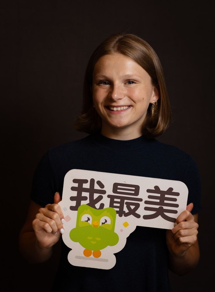 Smiling photo of Kayle — in a black blouse and bob — holding a green and white Duolingo sign with white fingernail polish
