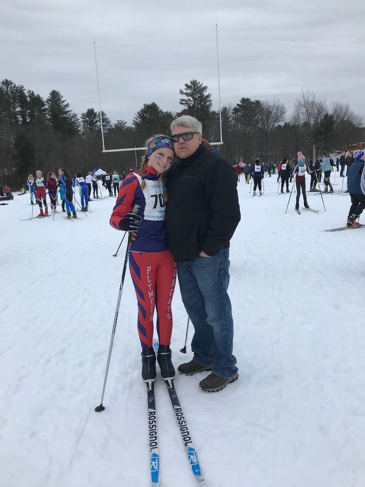Vivid, picturesque photo of a smiling Kaylie on snowy mountain top — in a red and blue ski suit and skis — hugging her Dad in a black jacket, black sunglasses and jeans after a high school ski competition 