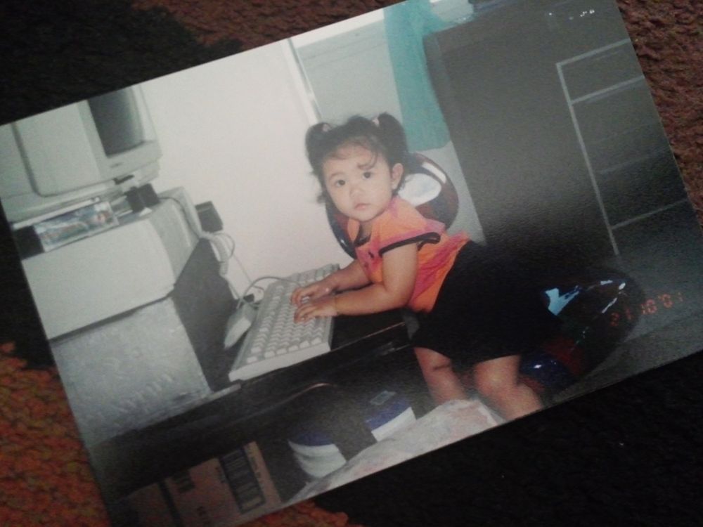 Color photo of a toddler Ashley Marie — in a pink and orange top and black shorts — looking toward the viewer while typing at a white desktop computer
