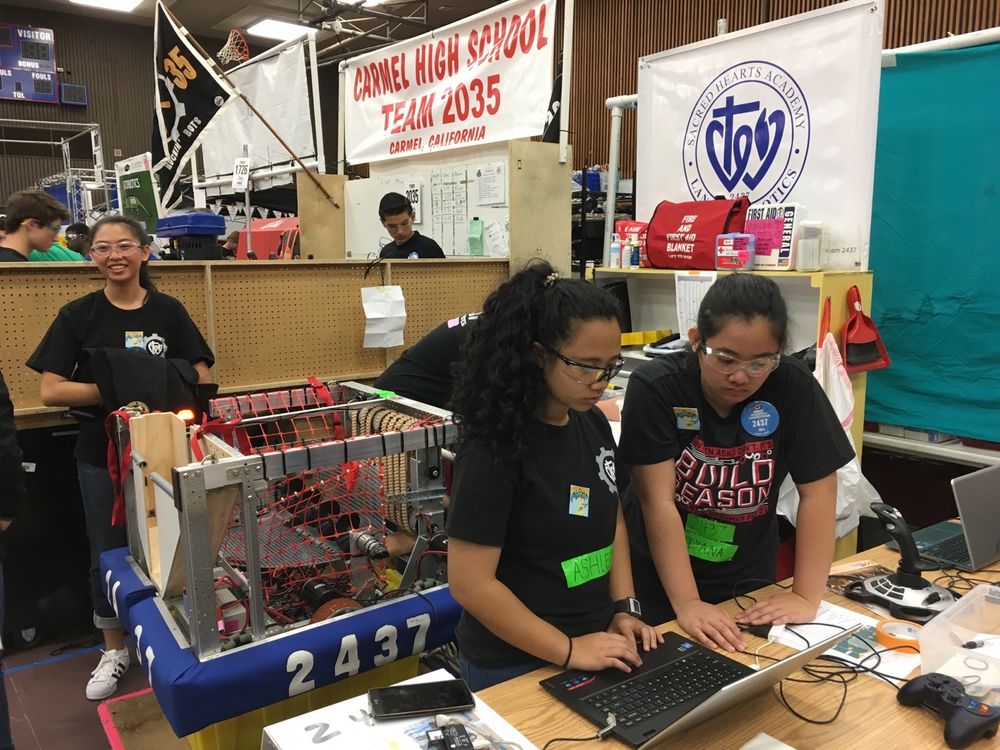 Color photo of Ashley Marie, wearing a black shirt and lab glasses, typing on a computer and working alongside her robotics teammates at a robotics competition 