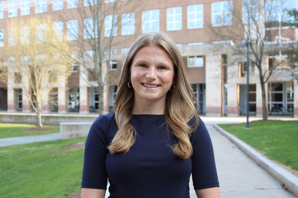 Smiling photo of Kaylie — in a navy blouse with curled hair and small gold hoops — standing in front of a building on Dartmouth’s campus