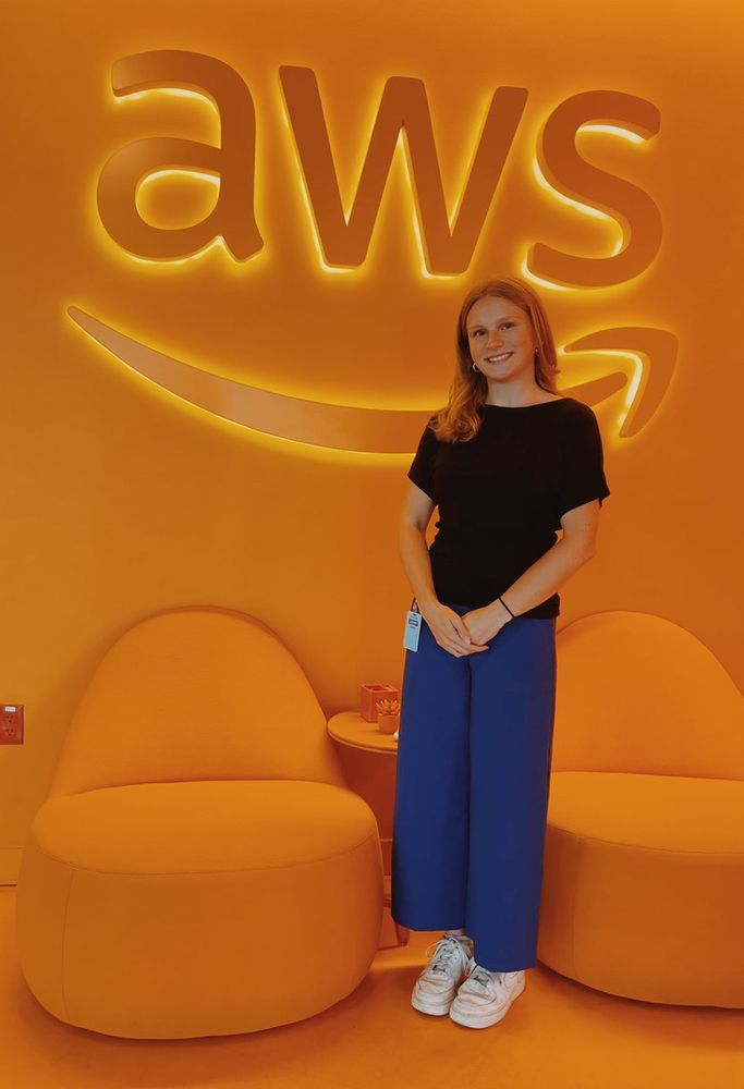 Vivid photo of Kaylie — in a black blouse and blue pants with white sneakers — standing in front of an orange AWS backdrop and next to two chairs