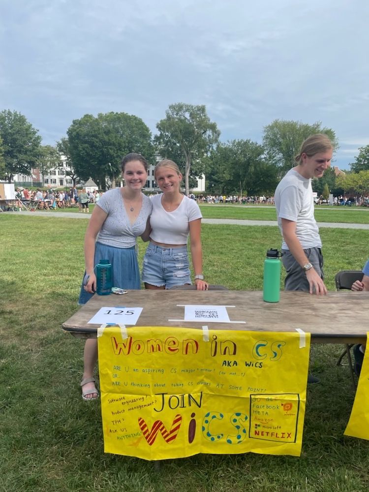 Color photo of Kaylie (R in blue shorts) and her colleague (L in a blue skirt)  from Women in CS club fair standing in front of a yellow banner and brown table at a student organization fair