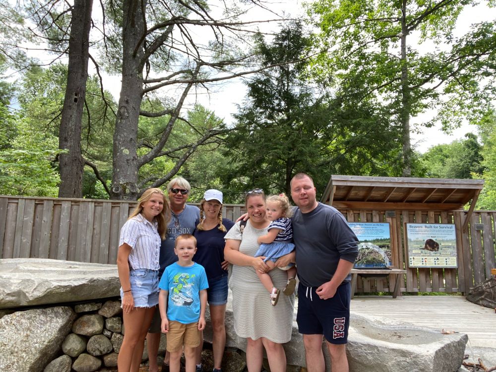Color group photo of Kaylie (L in jean shorts) with her Dad — in black sunglasses and a blue Red Sox shirt — hugging her Mom — in a navy shirt and white Nike hat — and four additional family members standing in front of trees
