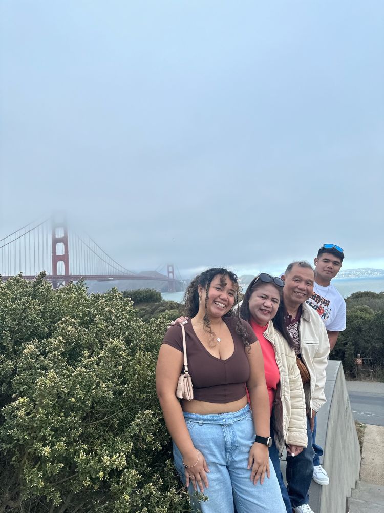 Color group photo of Ashley Marie (front in brown) with her mom (in a coral shirt and cream jacket), dad (in a brown jacket), and brother (in the back wearing a white t-shirt and sunglasses) standing in front of the Golden Gate Bridge in San Francisco