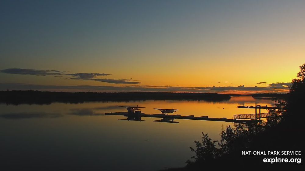 The golden sun sets over the Naknek River. Two float planes are parked at a floating dock. 