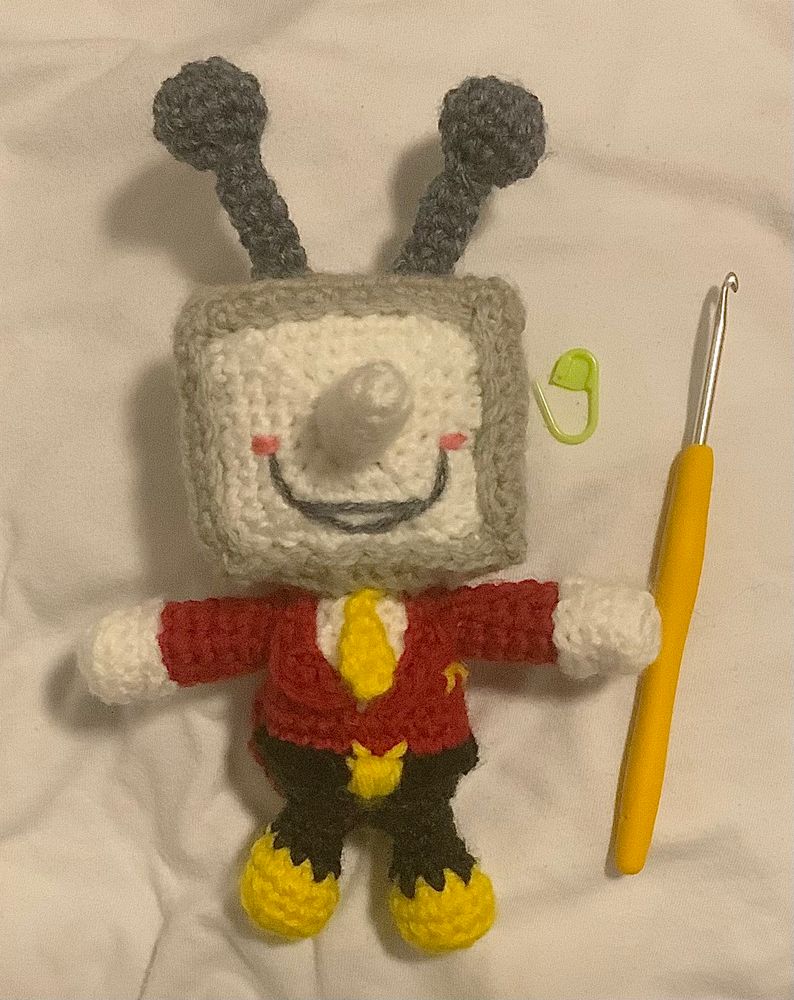 A front-facing view of the mini crochet doll of Tenna (from Deltarune) that I made! A cute little tv head man with a long nose, goofy little smile, and no visible eyes. He’s in a red suit with coattails, white gloves, yellow tie, black pants, and yellow shoes. He also has a belt and a yellow belt buckle embroidered to be shaped like his head (a tv with antennae)
He’s posed next to my crochet hook and one of my stitch markers!