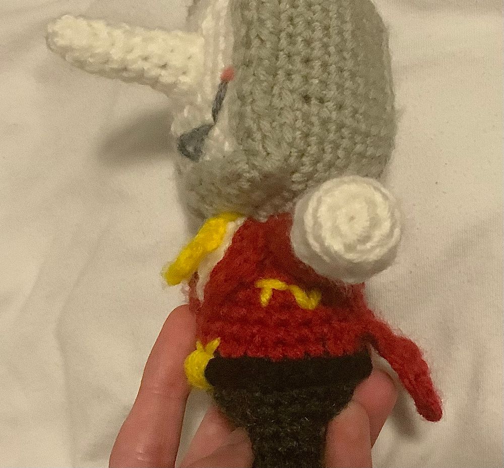 A close-up side view of the mini crochet doll of Tenna (from Deltarune) that I made! I lifted up his arm a little here to give a better look at the “TV” letters I embroidered in yellow on his suit