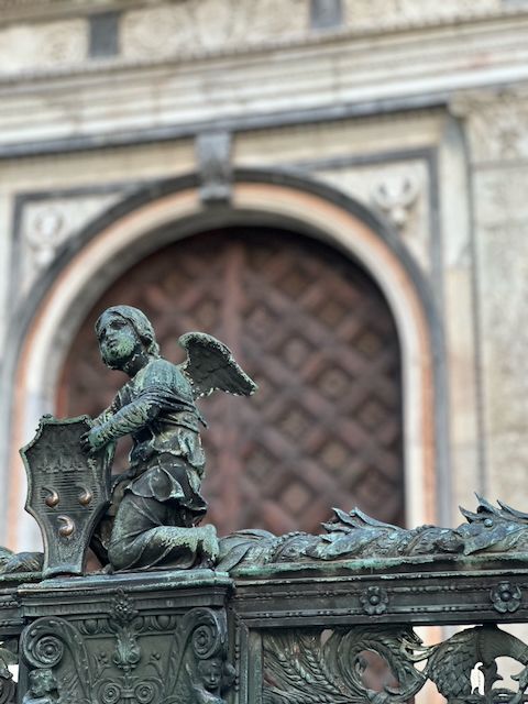 Bronze angel detail on the top of the elaborate Renaissance gate surrounding the Cappella Colleoni in the Città Alta in Bergamo, Italy.  The angel is kneeling and holding up a coat of arms plaque emballzoned with 3 testicles, which kind of look like 3 commas. These stylized testes are shiny bronze from people rubbing them (for luck?) while the rest of the gate's metal work is black but covered in beautiful verdigris deposits.  I have read that the tri-orchis coat of arms precedes the date of the chapel by at least several hundred years.

In the out of focus background are the arched carved wooden doors to this Bartolomeo Colleoni funerary chapel built in 1486, with the surrounding wall being made of highly detailed carved and adorned pale cream and grey marble.  There are various additional testicular coats of arms on both the gate top (in bronze) and in marble on the walls of the building.  That's a lotta balls.
