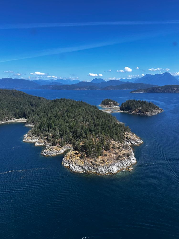 Aerial photo of the vast British Columbia island landscape showing blue waters of the Salish Sea and various heavily wooded rocky islands in the foreground and background. there are a few perfect puffy white clouds in the background over the mountains. Two streaks across the upper horizon appeared to be contrails from jets, but are actually distortion from the curved window I was photographing through. The perspective is from a low-altitude float plane.