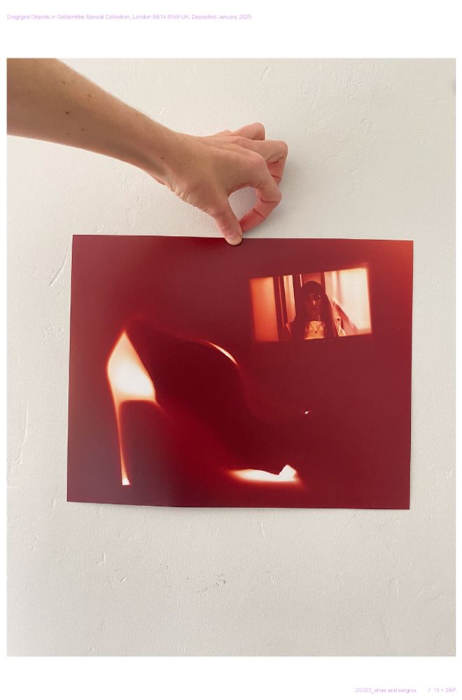 Red photogram  / contact print of a still from an online performance by #Sergina from 2020 and a high heeled shoe. The print is held up against a grey wall by a hand. 