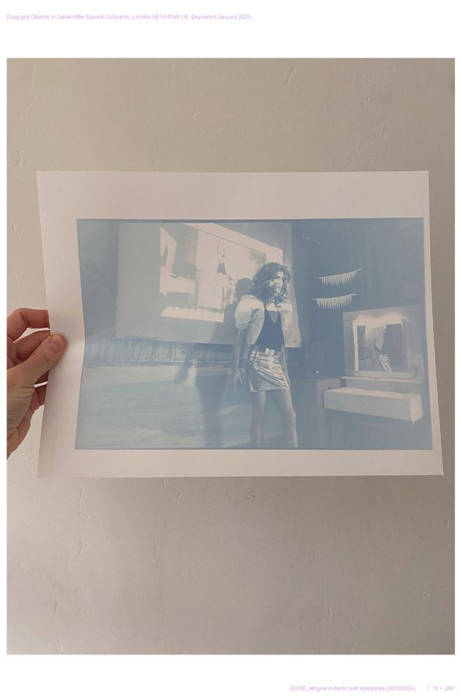 Hand printed photograph/photogram showing a drag queen standing patiently during the setting up of Google Hangout in the back of Silver Futures bar in Berlin in 2016. #Sergina is played here by Thomas Doherty. The imprint of two false eyelashes that were laid on the photo paper at the time of printing hover beside the queen. The tone of the image is blue. In a mirror to the right a reflection of #Sergina appears. On the left of the image is a hand holding the image. 