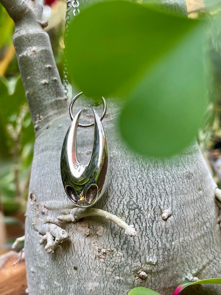 A shiny silver pendant, shaped like the stylized, horned shell of Hornet from Hollow Knight, hangs from a silver chain against the light gray, textured trunk of a plant. A large green leaf partially covers the top of the image.