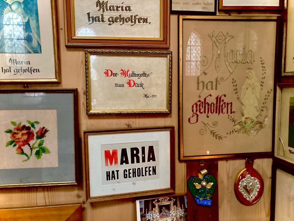 Maria hat geholfen.
Wand mit gestickten Bilderrahmen in einer Kirche.