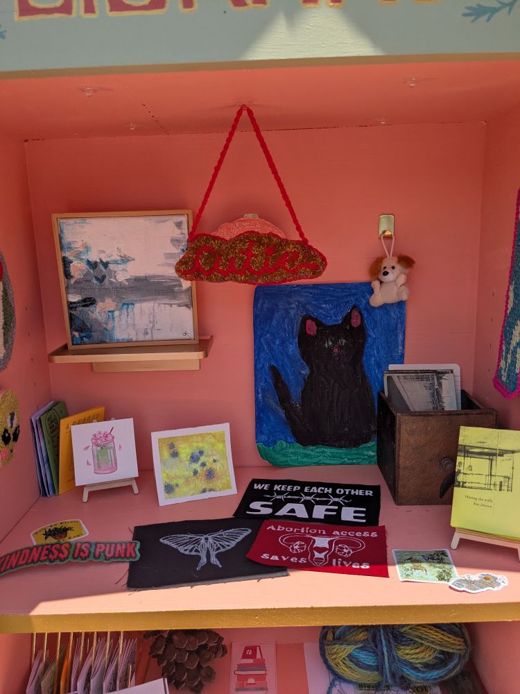The inside of the little free art library which is painted bright peach. On a small gold shelf, there's an abstract grey, blue, and peach painting in a wooden frame. There's a large painting of a big black cat against a vibrant blue sky. There's an old wood box full of old postcards and a tiny watercolor painting of a matcha tea. There's a bunch of block prints on fabric - one says "we keep each other safe", another has a Luna moth, and another says "abortion access saves lives". Hanging from the top is a punch needle piece which says "cutie". Come touch our art if you like!