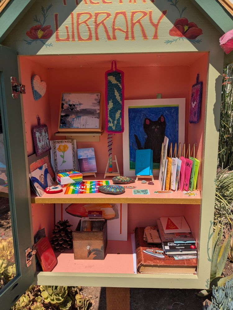 The little free library which is sage green outside and bright peach inside. It's full of art! There's a picture of a big black cat on a blue background, a Tunisian hanging graphgan of leaves on a teal background, sage wrapped in brightly colored yarn, a zine with a poppy on the cover, a rainbow colored fabric patch that says "queer" with barbed wire around it in a heart, a trans pride heart punch needle pin, and lots more stuff.
