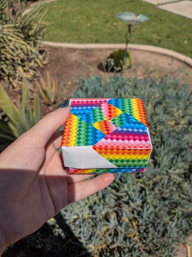 A small origami box made of rainbow colored paper. It has a lid that you can take off.