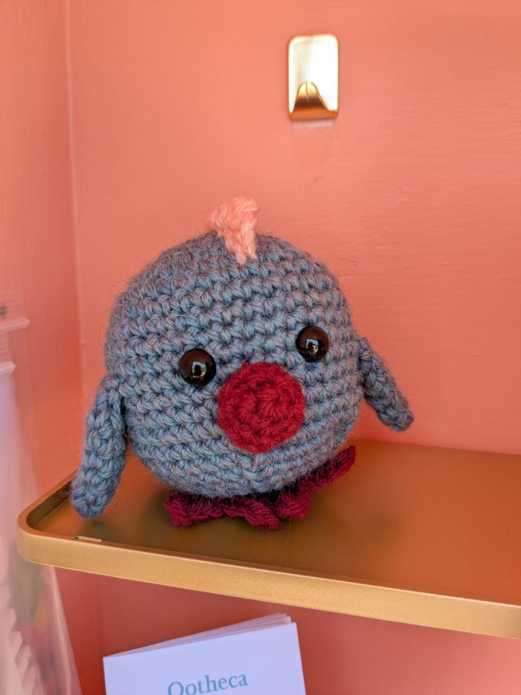 A little crochet chick that is a perfect ball. It's body is a variegated blue wool yarn and it's beak and feet are a dark terracotta red. It's comb is peachy pink and he has two black plastic eyes. You are welcome to touch anything in the library!