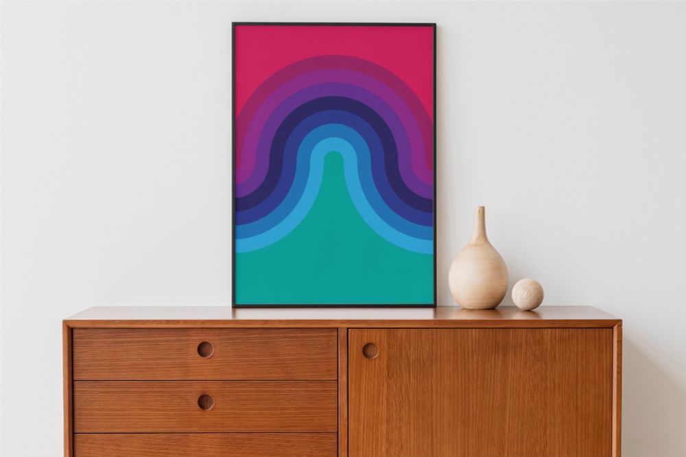 A colorful framed artwork displayed on a wooden credenza. The design features a series of concentric arcs in shades of purple, blue, and teal, set against a red background. Two wooden decorative objects—a vase and a sphere—sit on the credenza to the right of the frame.