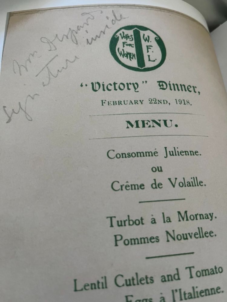 Photo of a menu from the book "Menu". Green writing says "Victory Dinner, Feburary 22ndf, 1918, and a pescatarian menu written in French. On the top left side in cursive handwriting are the words "Mrs Despard's signature inside"