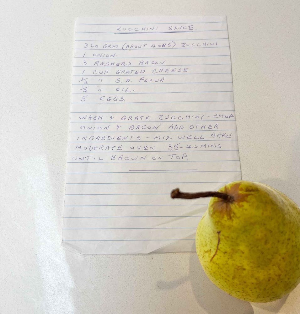 A handwritten recipe and a pear. The recipe reads: 
Zucchini slice
360 gram (about 4 or 5) zucchini
1 onion
3 rashers bacon
1 cup grated cheese
1/2 cup s.r. flour
1/2 cup oil
5 eggs
Wash and grate zucchini. Chop onion and bacon. Add other ingredients. Mix well. Bake moderate oven 35-40 mins until brown on top. 

Note: Google says "A moderate oven typically refers to a temperature range of 177-191°C (350-375°F)."