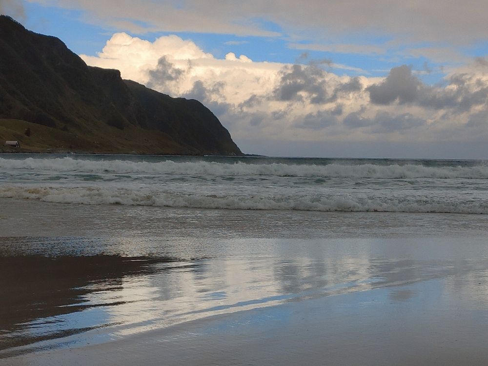 Man sieht einen steilen Bergabhang am Meer, in Hintergrund weiße und graue Wolken. Unten ist der leicht überflutete Strand, in dem sich die ganze Szene spiegelt. Man könnte das Bild auch verkehrt rum halten.