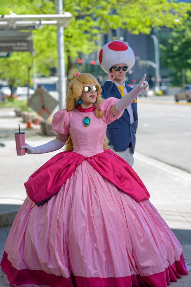 A cosplay of Princess Peach. She has heart sunglasses on, is pointing at something out of frame, and a man with a Toad hat in the back as a bodyguard