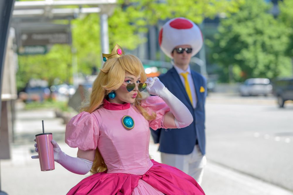 A cosplay of Princess Peach. She has heart sunglasses on and a man with a Toad hat in the back as a bodyguard. 