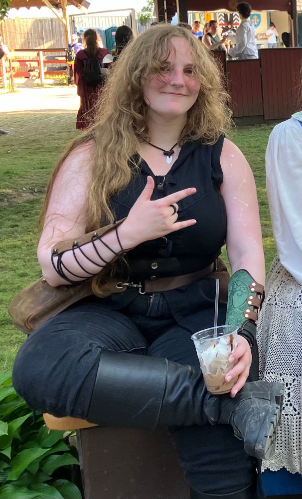 An image of Red at the renfaire, dressed in a mostly-black roguish ensemble with no sleeves. The constellation designs are visible on her left arm. She's also holding a very period-accurate iced cappuccino.
