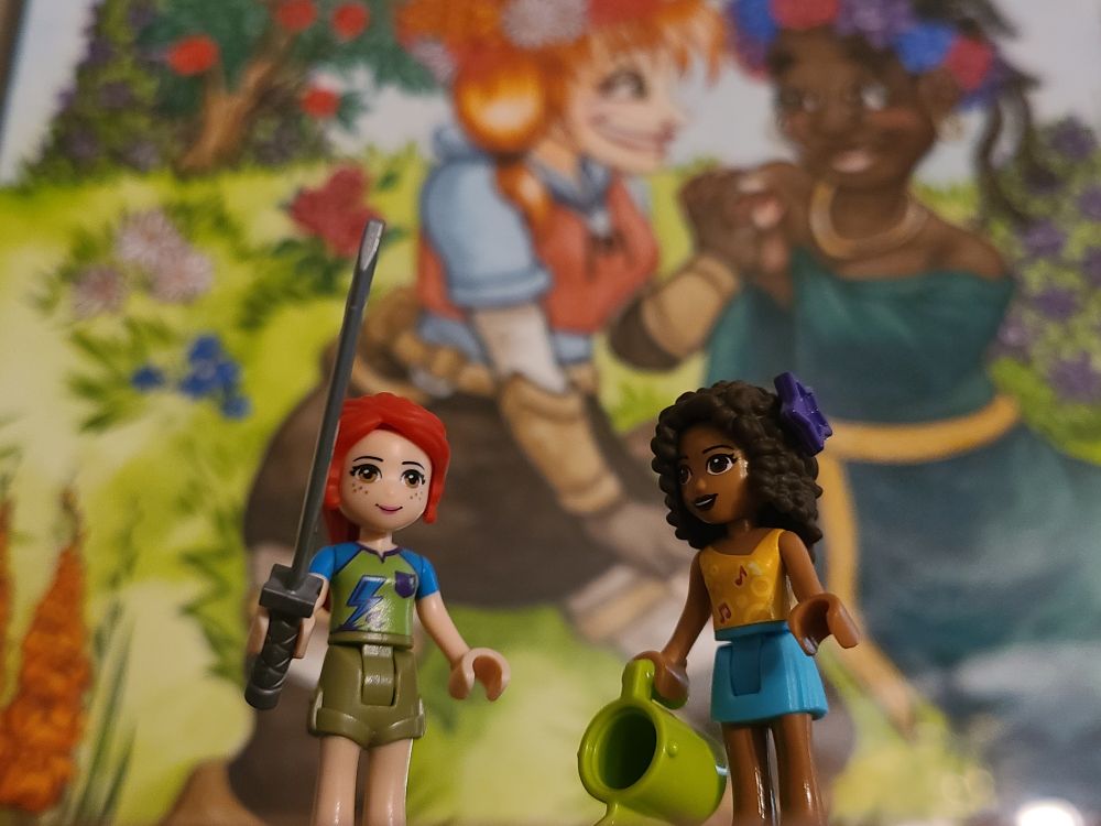 Lego Minifigs that look like OC's in front of commissioned art of them. A redheaded white girl with freckles holding a sword and a Black girl with natural curls holding a watering can.