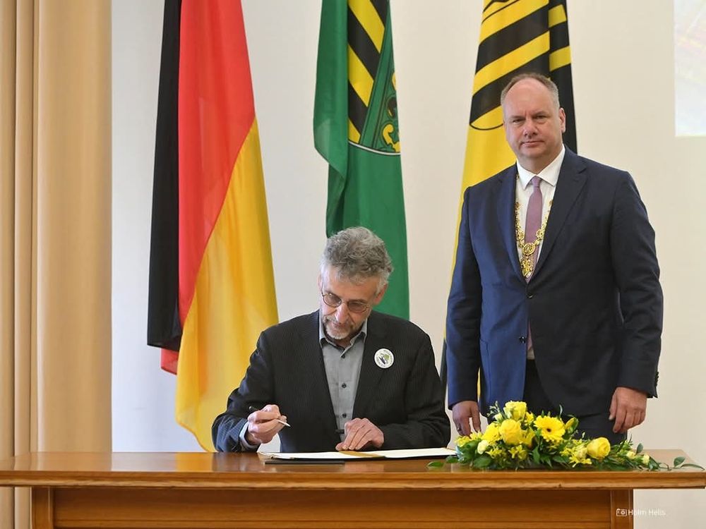 Sitzend Michael Kobel beim Eintrag ins Preisträgerbuch mit Button von Willkommen in Löbtau am Revers. Stehend dahinter OB Hilbert.
Im Hintergrund die Fahnen von Deutschland, Sachsen und Dresden.