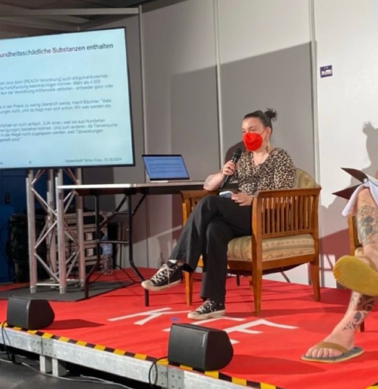 a person wearing a red ffp2 mask is talking into a microphone while sitting on a chair on a stage. to their right is a screen with a presentation, just out of frame to their left is another person also sitting in a chair