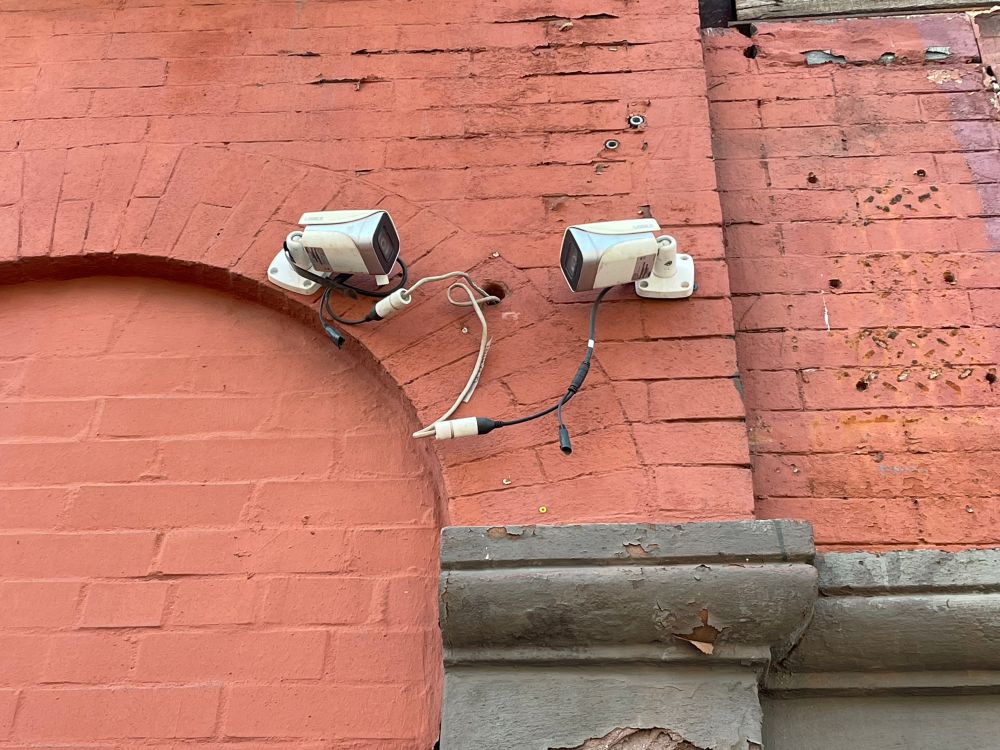 Two security cameras very close together and facing each other on the exterior of a brick apartment building.