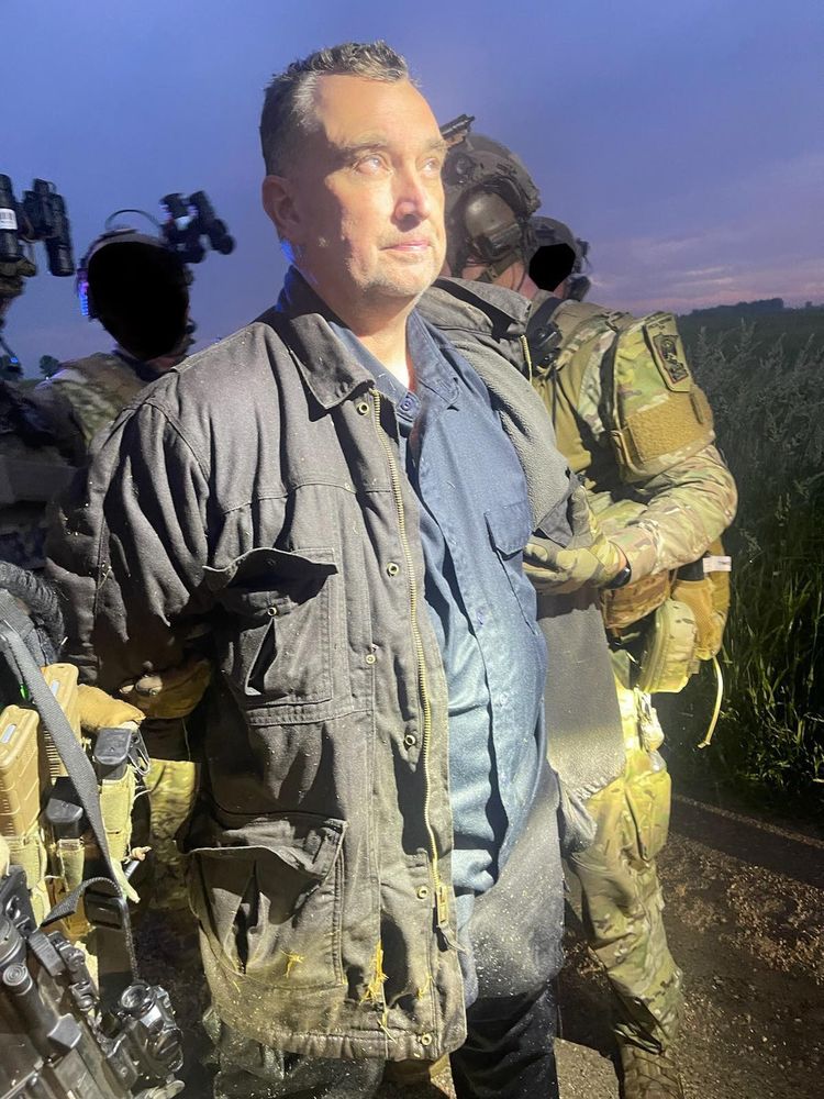 Slightly eerie photo of a white man wearing a dark blue shirt and green jacket being taken into custody by officers in camo. The man has bright lights on his face, there's a field behind him, and the sky is twilit.
