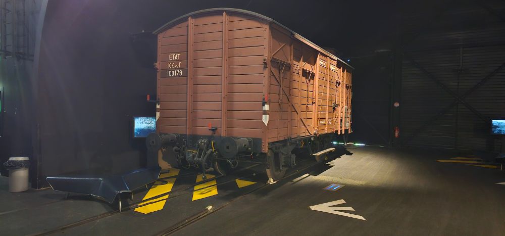 Un wagon à bestiaux dans un hangar sombre, avec une petite télé sur la gauche, un peu cachée. 