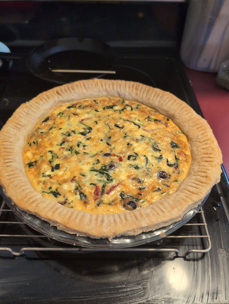 A home made quiche with fork pressed edge pie crust. Spinach, roasted red peppers, kalmata olives, and sundried tomatoes can be seen in the filling