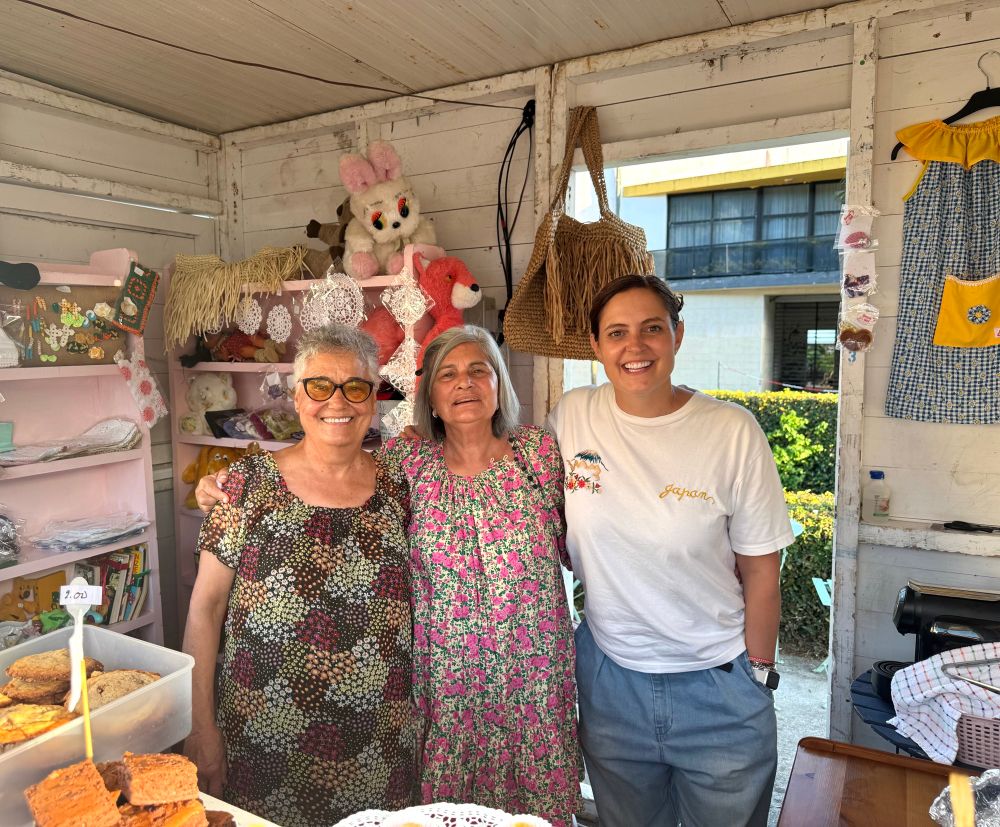 In einer Marktbude auf einem Fest in Horta stehen drei Frauen unterschiedlichen Alters, die alle für „Dress a Girl“ tätig sind. Drumherum selbstgebackene Kuchen und Bekleidung zum Verkauf fürcdie Organisation.