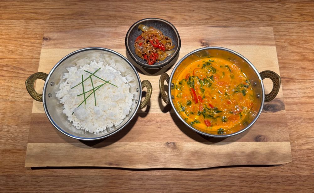 Auf einem Holzbrettchen stehen zwei indische Servierschalen, eine (links) ist mit dampfendem Basmati-Reis gefüllt, darüber Schnittlauch-Stengel, rechts die Schale ist mit rot-orangener Sauce gefüllt, in der der grüne Blattspinat Farb-Akzente gibt. In der Mitte oberhalb der Schalen ist eine kleine schwarze Schale mit der Würzsauce, dunkelrot.