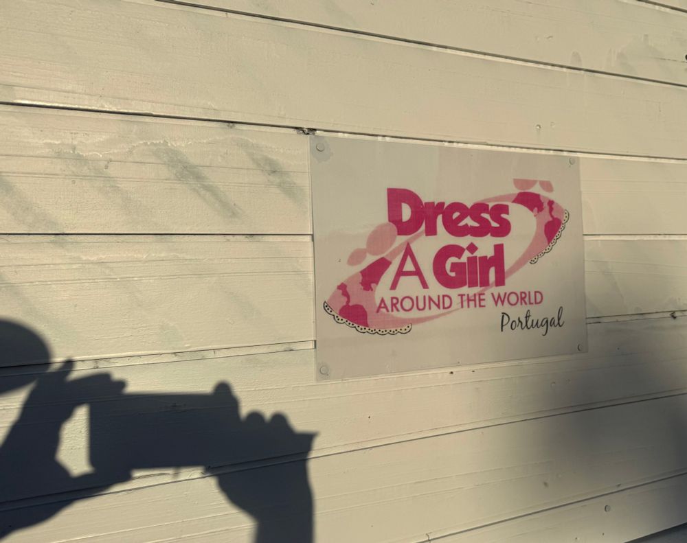 Logo der weltweit tätigen Organisation „Dress A Girl“ auf der Seitenwand der Bude.links davon mein Schatten beim Fotografieren.