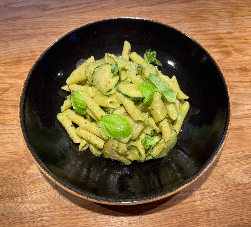 Schwarzer runder Teller mit Penne, die in cremiger hellgrüner Sauce verrührt sind. Einzelne Scheiben Zucchini zur Deko mit Basilikum-Blättern. 