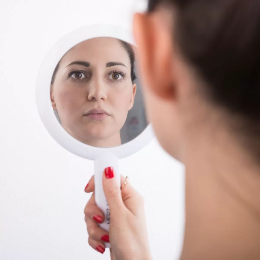 A woman viewing her own reflection in a hand-held mirror.