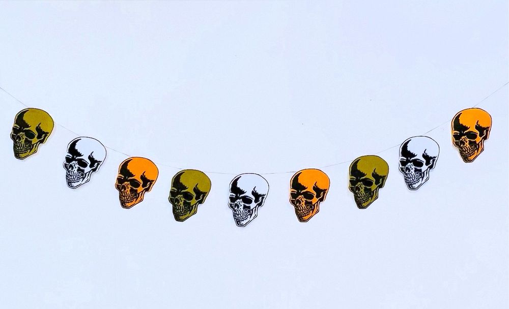 A garland banner of 9 linocut skulls printed in black, orange and green hanging in a swag shape against a white wall. 